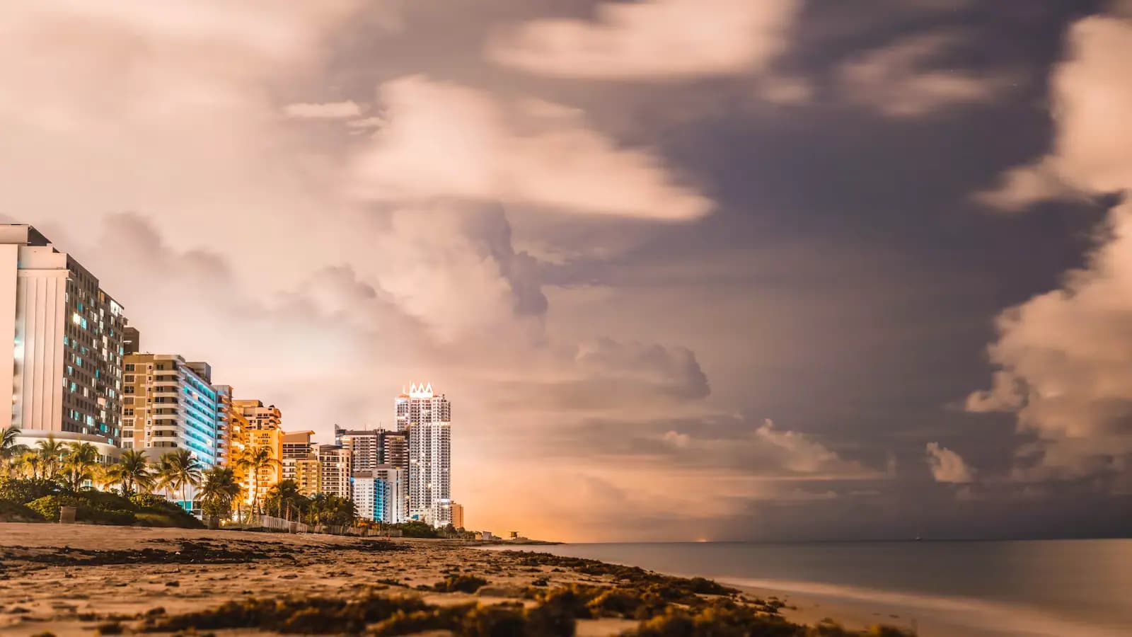 Storm clouds building over a barrier-island shoreline at dusk with lit hotel towers along the sand