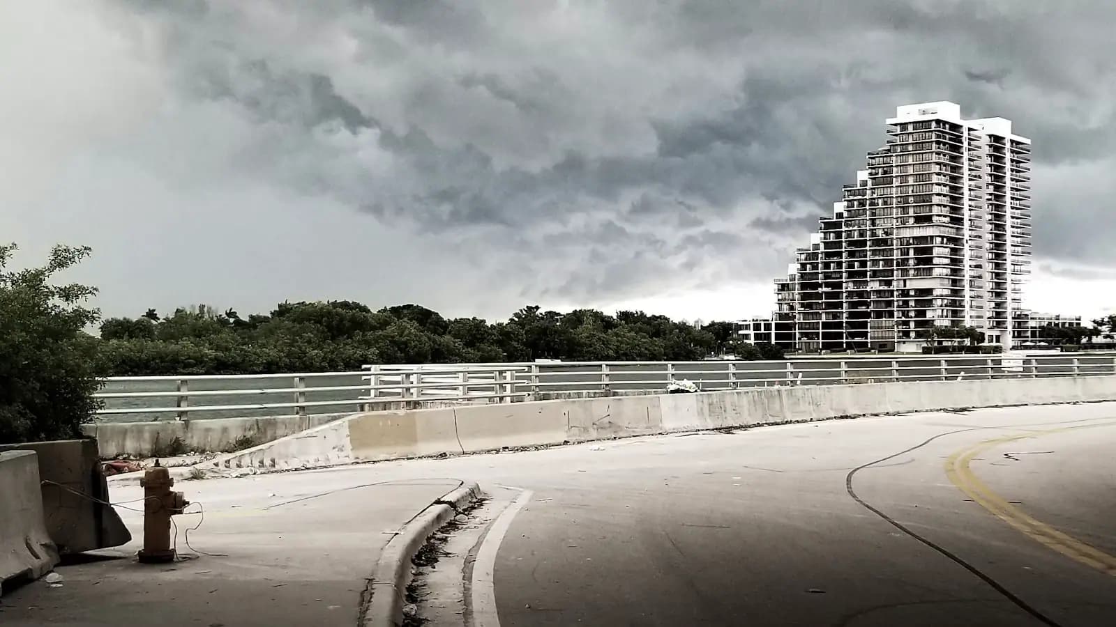 A causeway road with white railings curving towards a high-rise skyline under heavy grey cloud