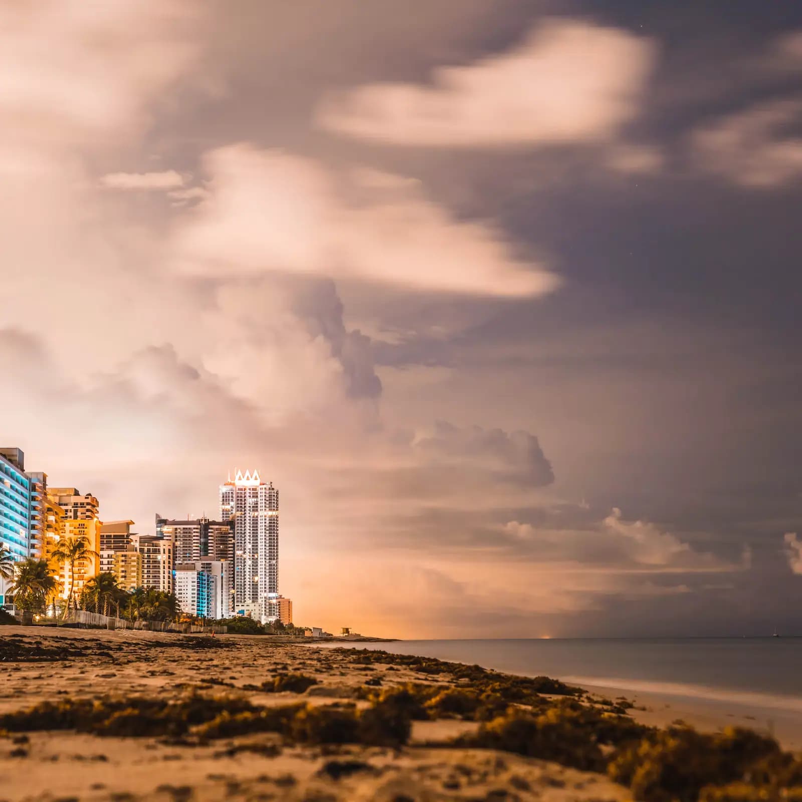 Storm clouds building over a barrier-island shoreline at dusk with lit hotel towers along the sand