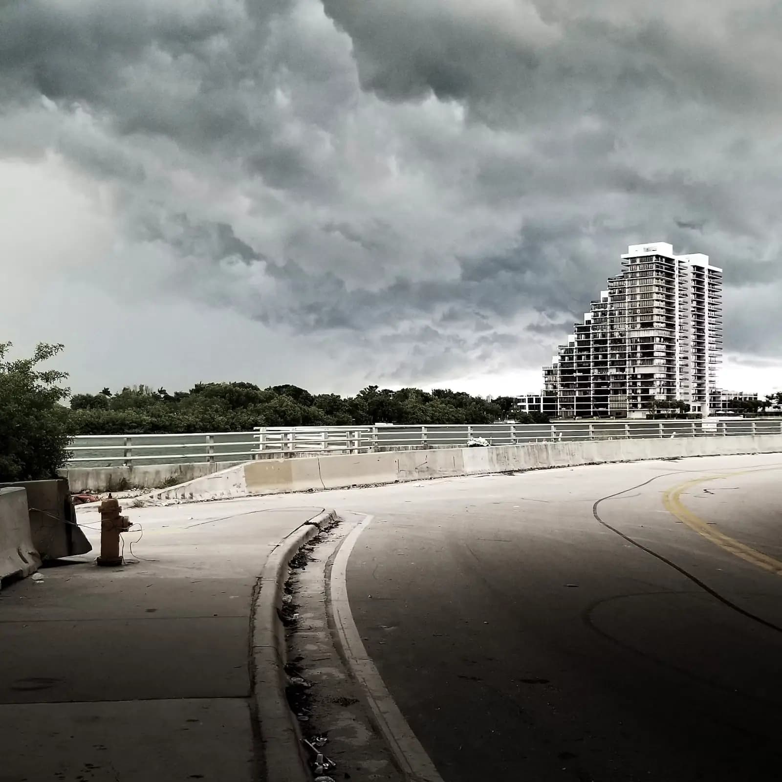 A causeway road with white railings curving towards a high-rise skyline under heavy grey cloud