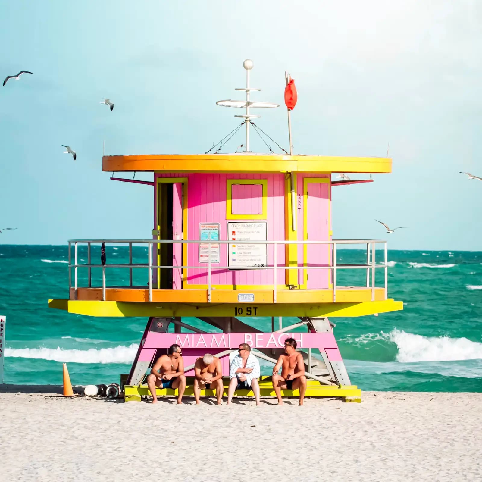 A pink and yellow lifeguard tower on South Beach with people sitting in the shade beneath it