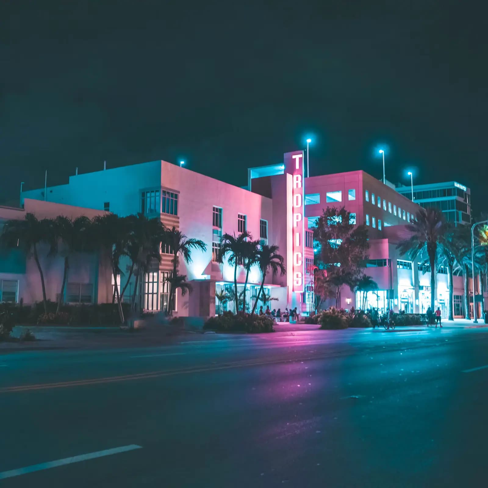 A neon-lit Art Deco hotel frontage with a vertical name sign on a palm-lined street after dark