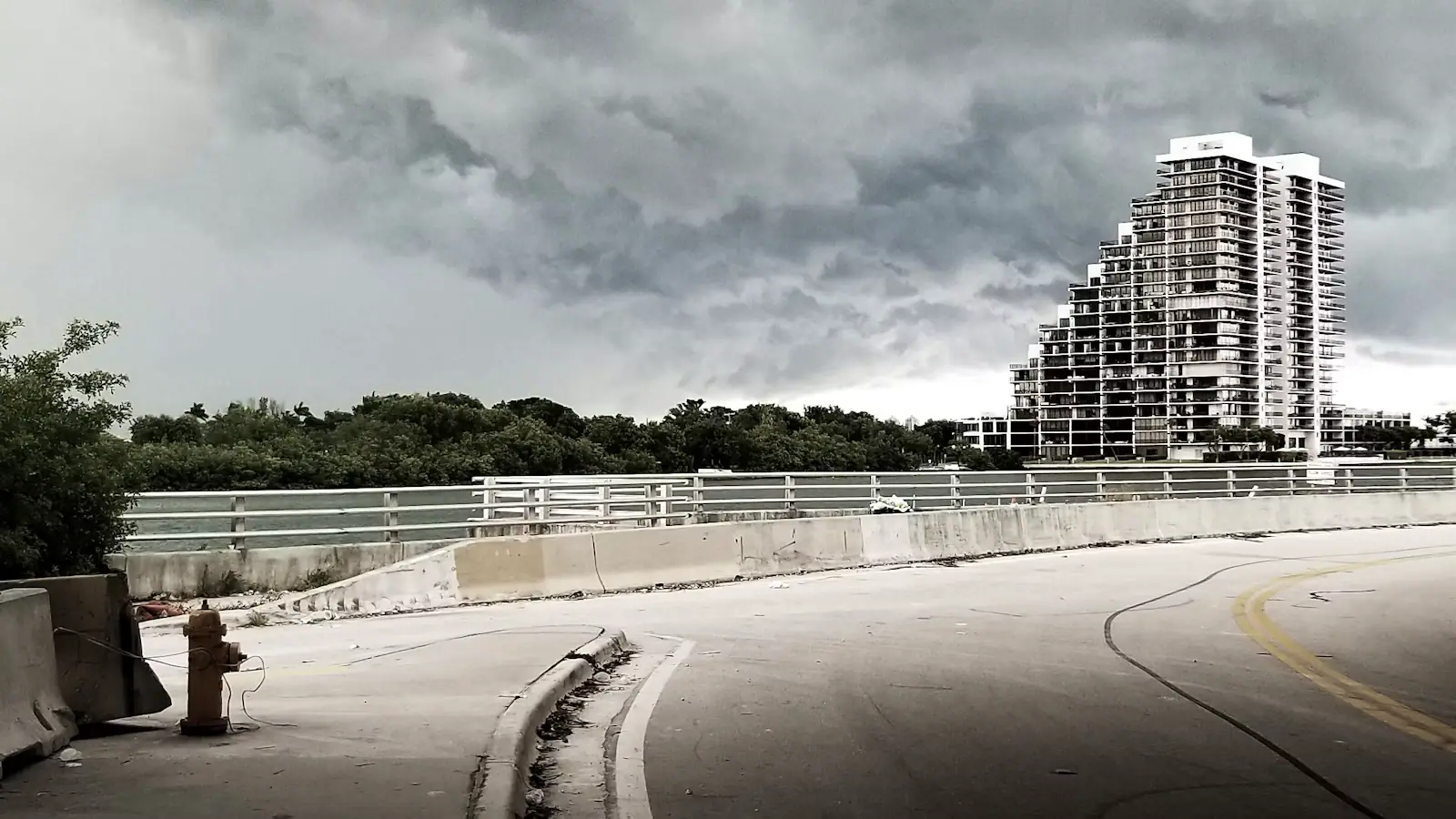 A causeway road with white railings curving towards a high-rise skyline under heavy grey cloud