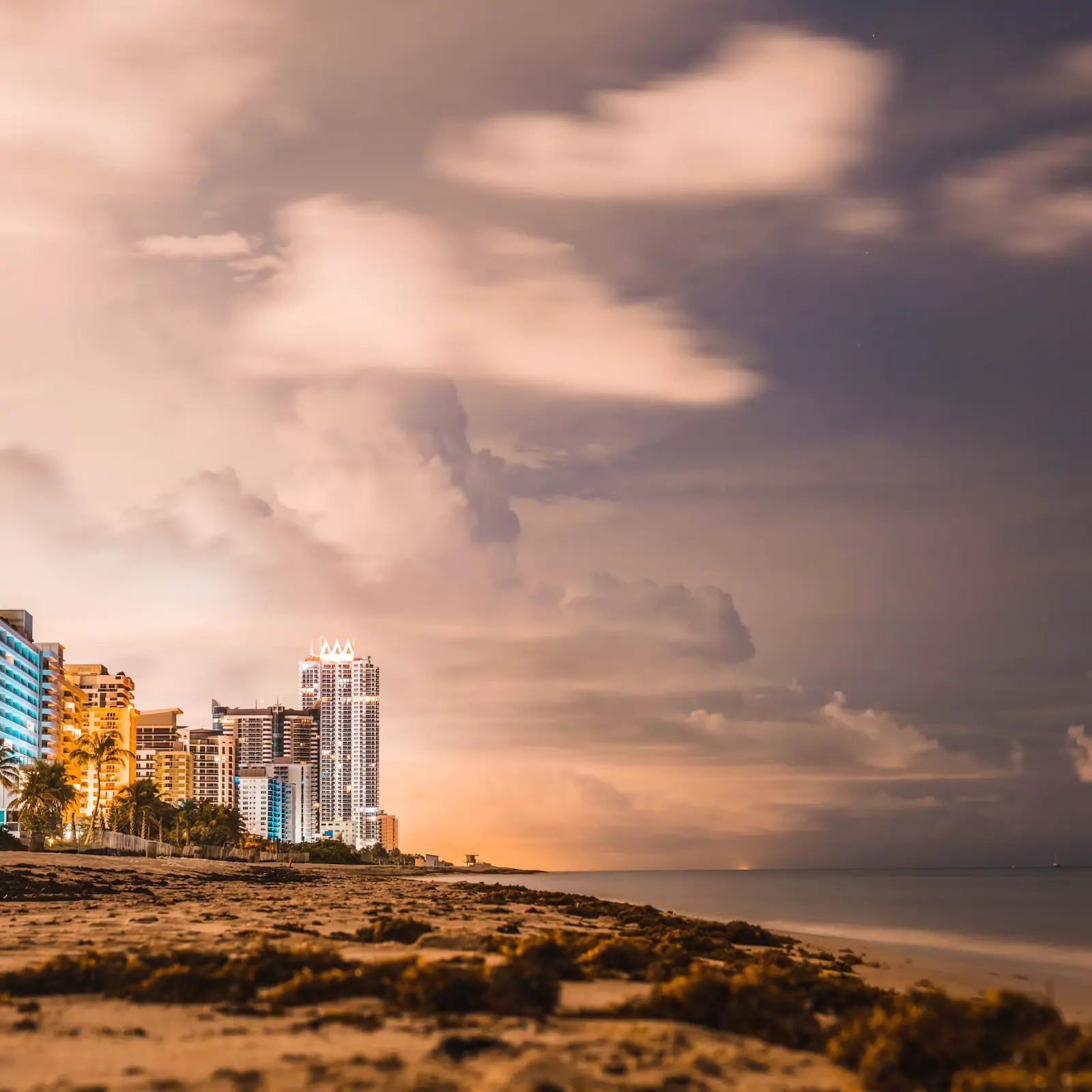 Storm clouds building over a barrier-island shoreline at dusk with lit hotel towers along the sand