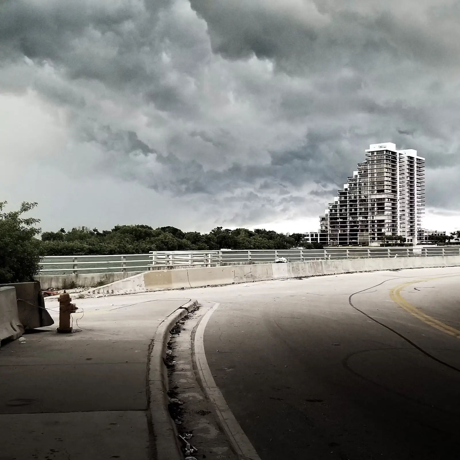 A causeway road with white railings curving towards a high-rise skyline under heavy grey cloud