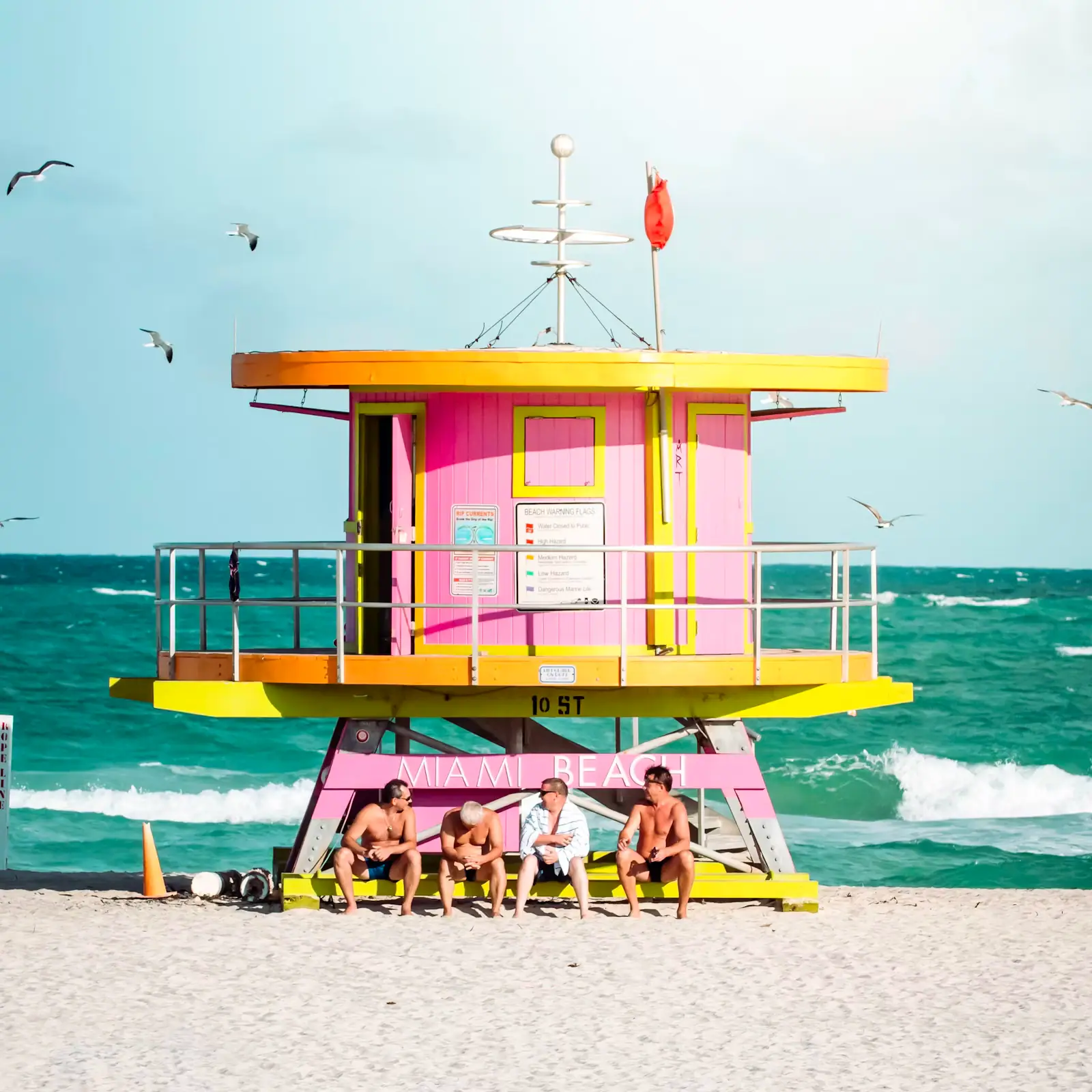 A pink and yellow lifeguard tower on South Beach with people sitting in the shade beneath it